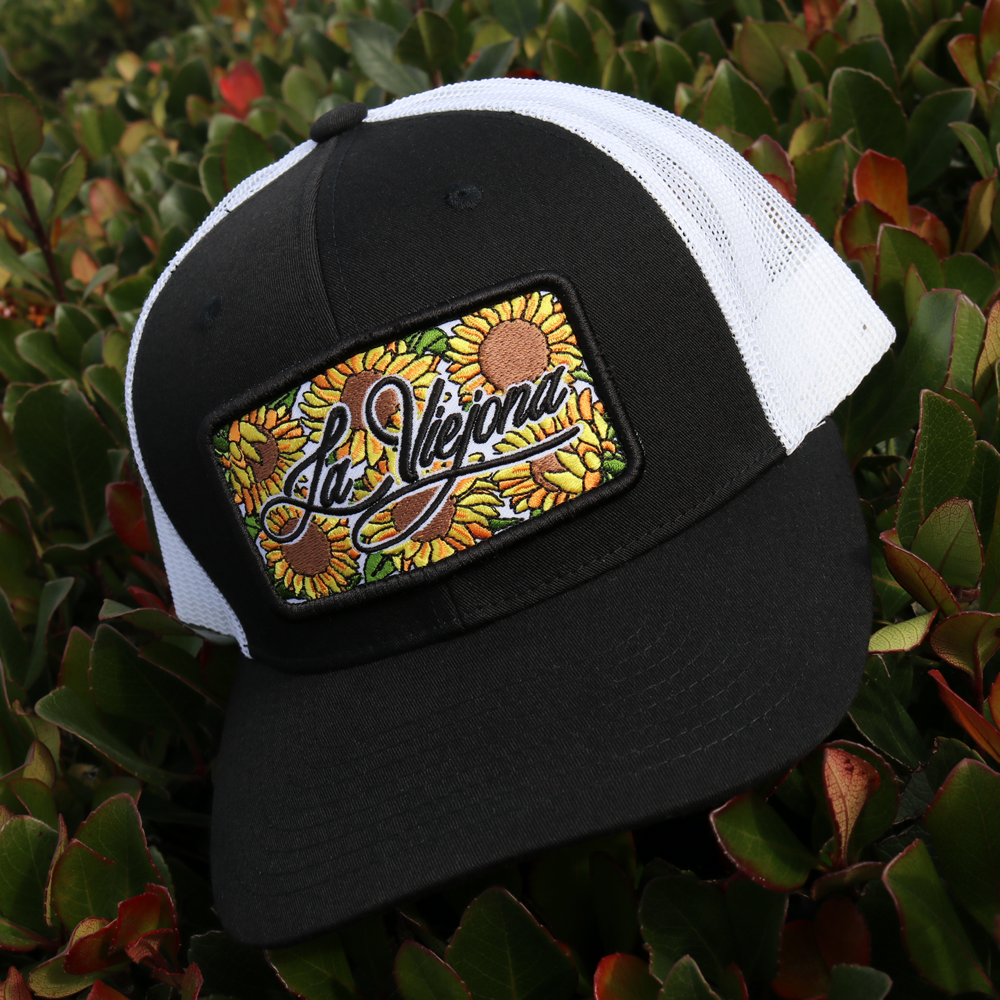 Black and white TruCap™ trucker hat with "La Vieja" sunflower patch, pre-curved visor, and mesh back.
