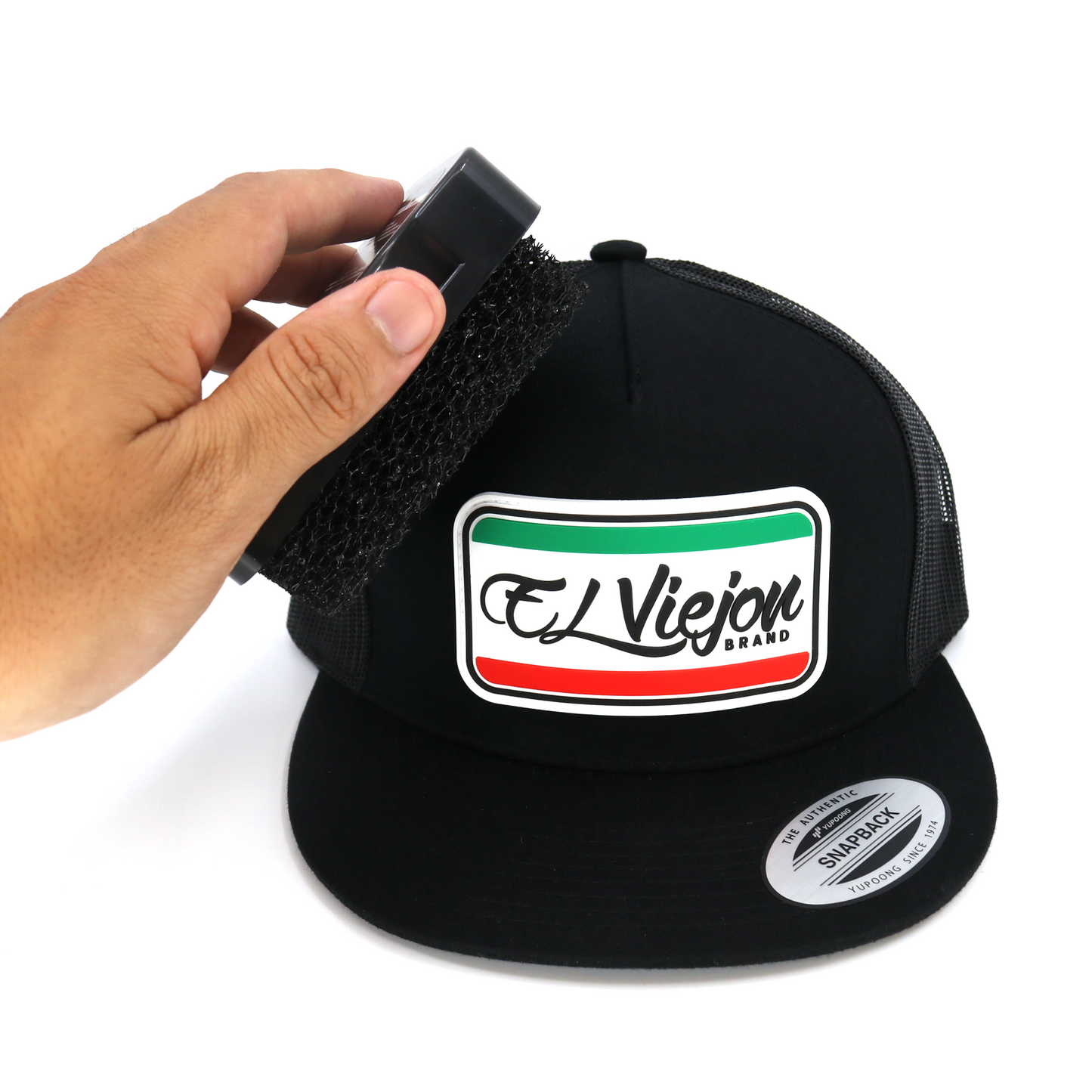 Hand using a black foam El Viejon hat brush to clean a black snapback hat.