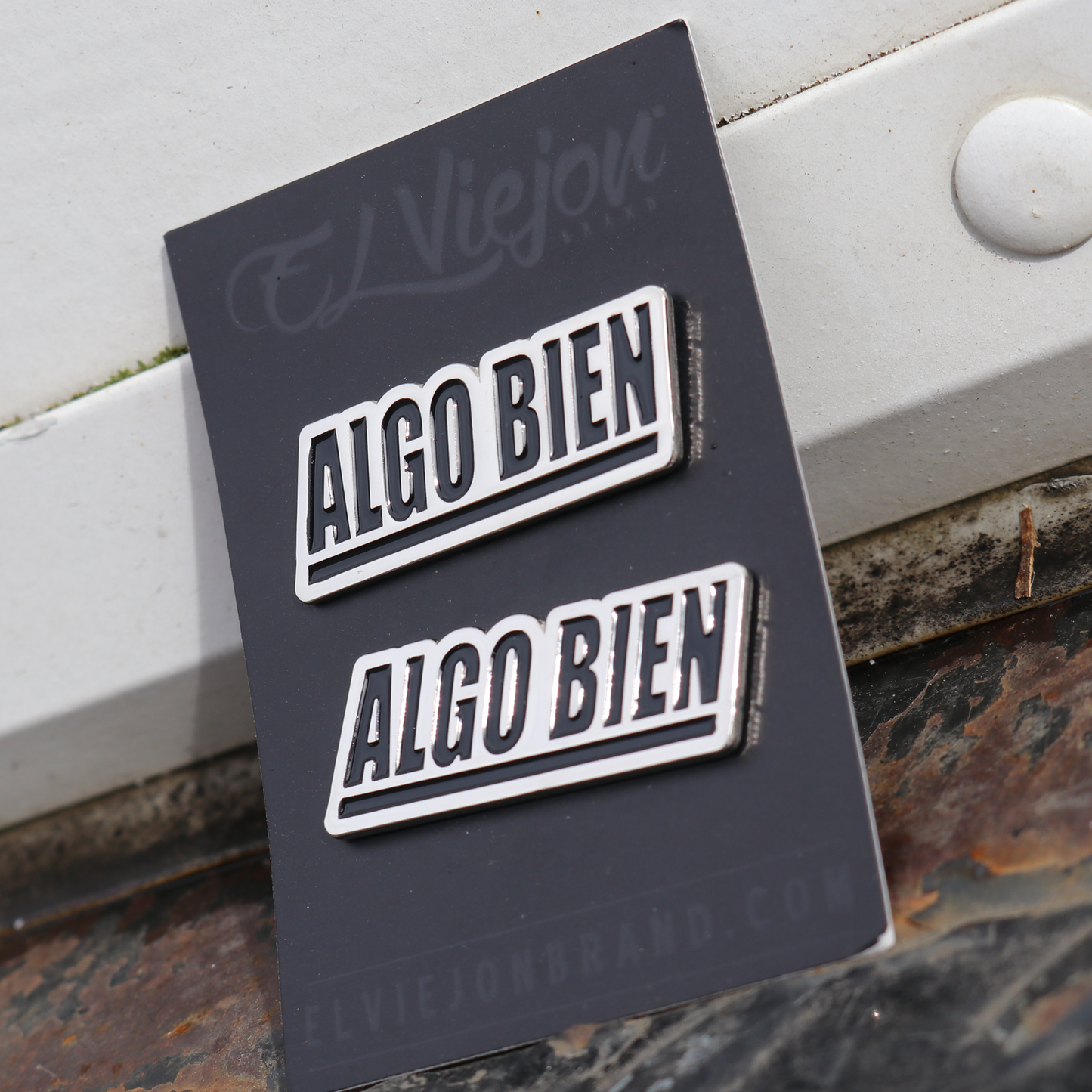 Two silver and black "ALGO BIEN" enamel pins on a dark gray El Viejon Brand card.