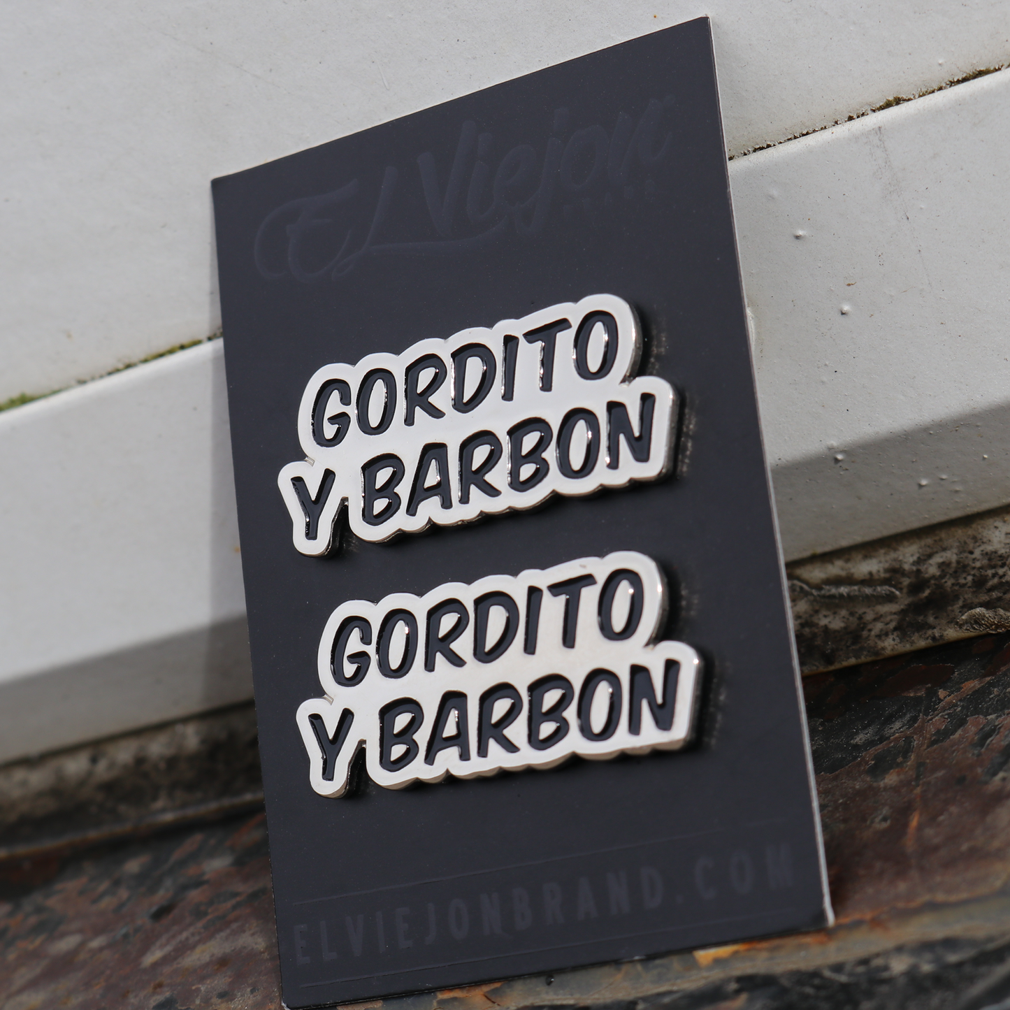 Two silver "GORDITO Y BARBON" metal pins with black outlines on a black display card.