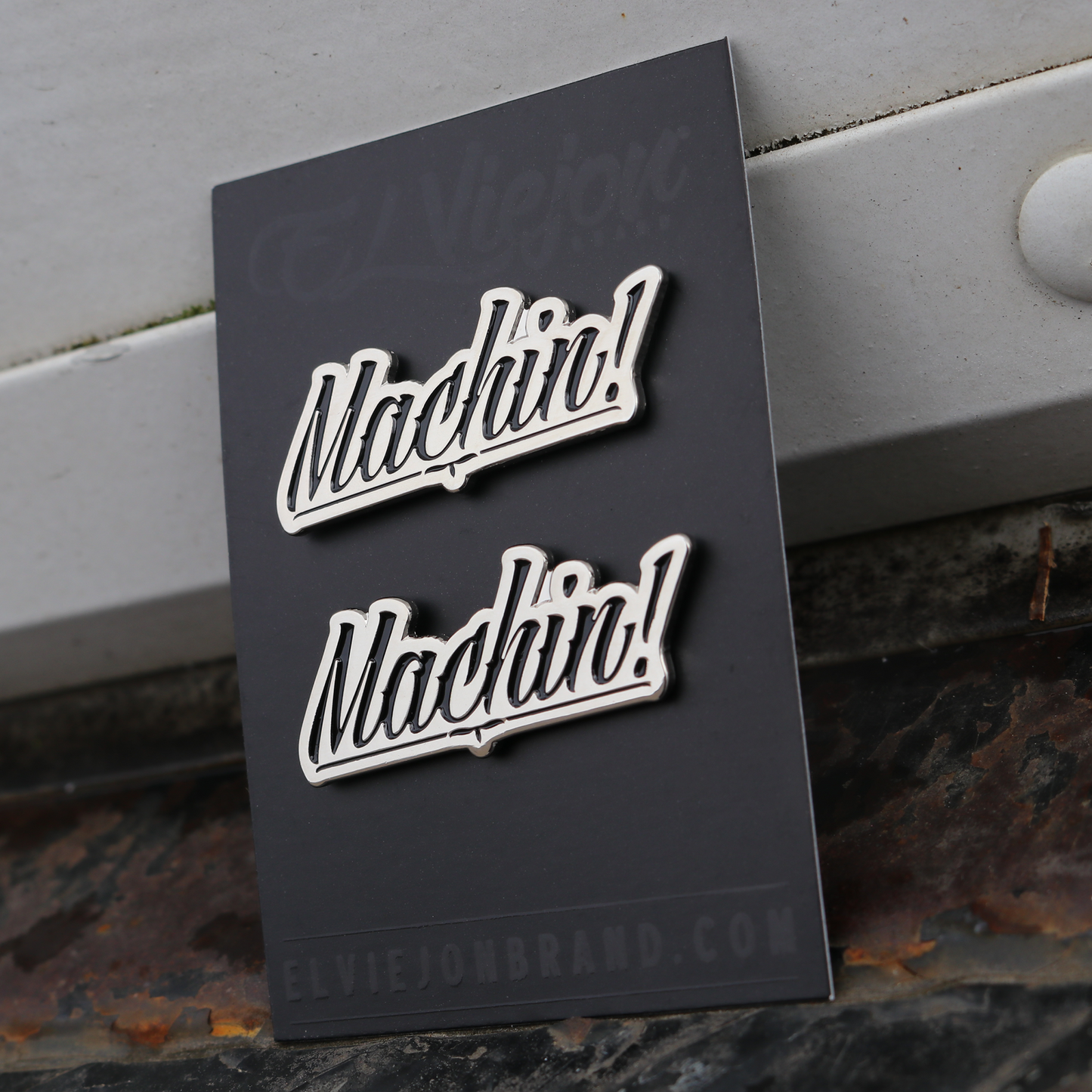 Two silver Machin! script pins on a black card, with rubber clutches.