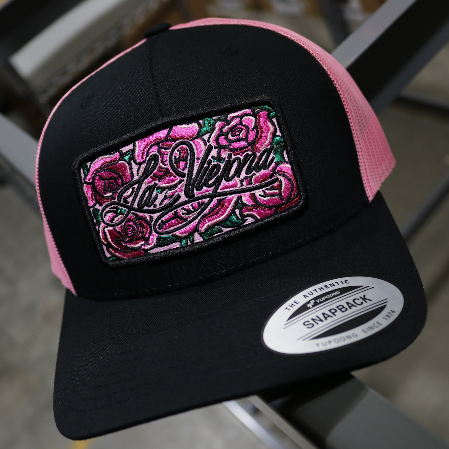 Black Yupoong trucker hat with hot pink mesh, embroidered "La Viejona" roses patch, and snapback.