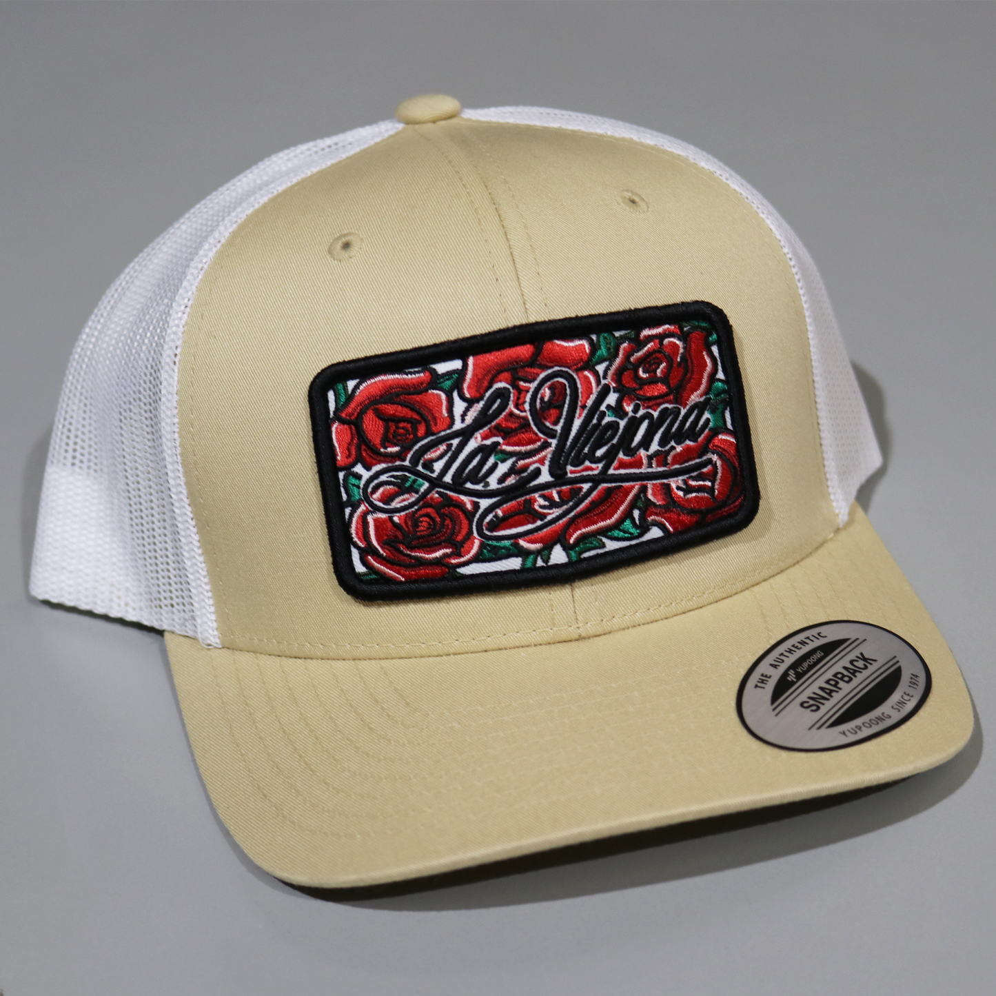 Khaki and white Yupoong Classic trucker hat with a black patch featuring red roses and "La Viejona" script.