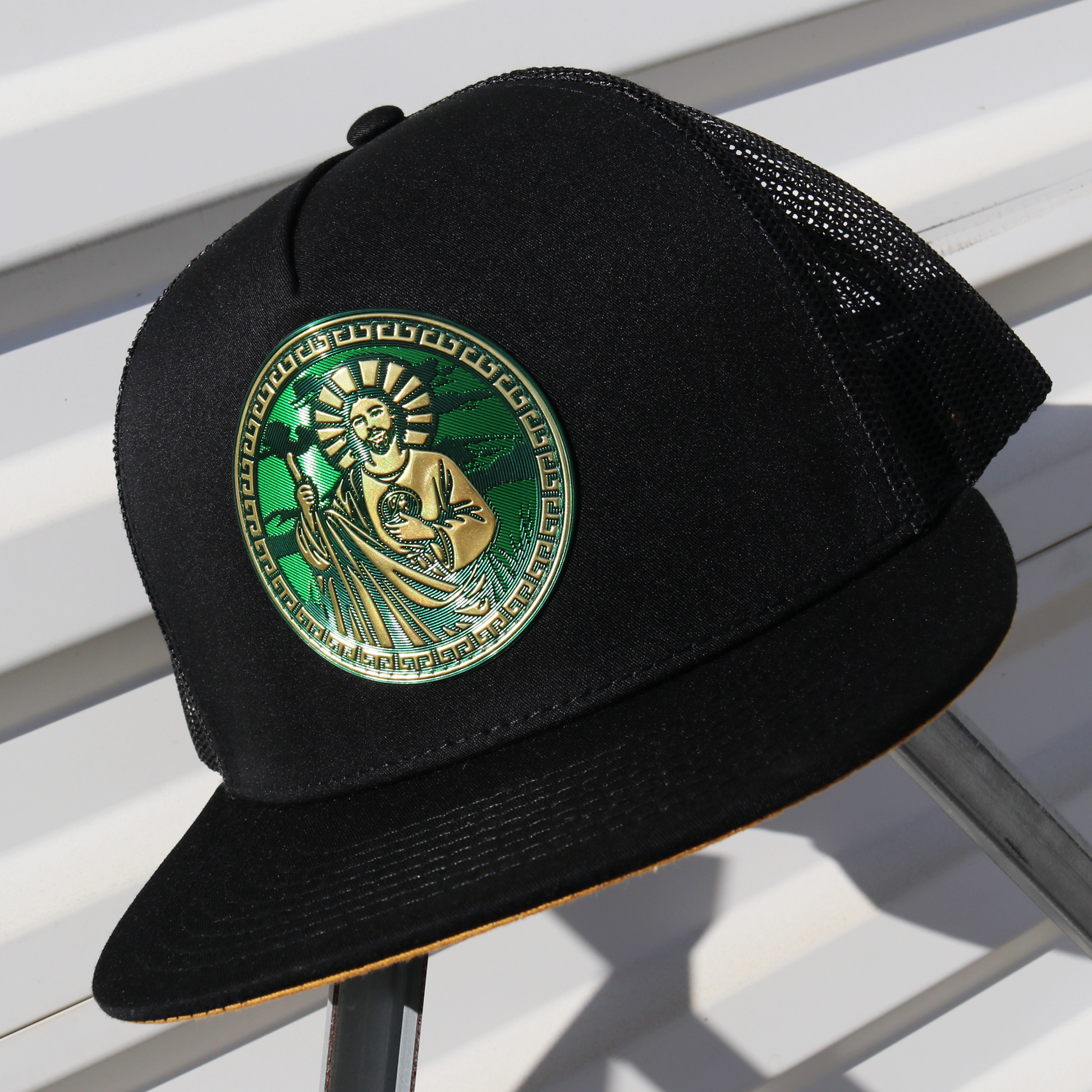 San Judas black flat visor trucker hat with green and gold emblem, black mesh back, and adjustable snapback.