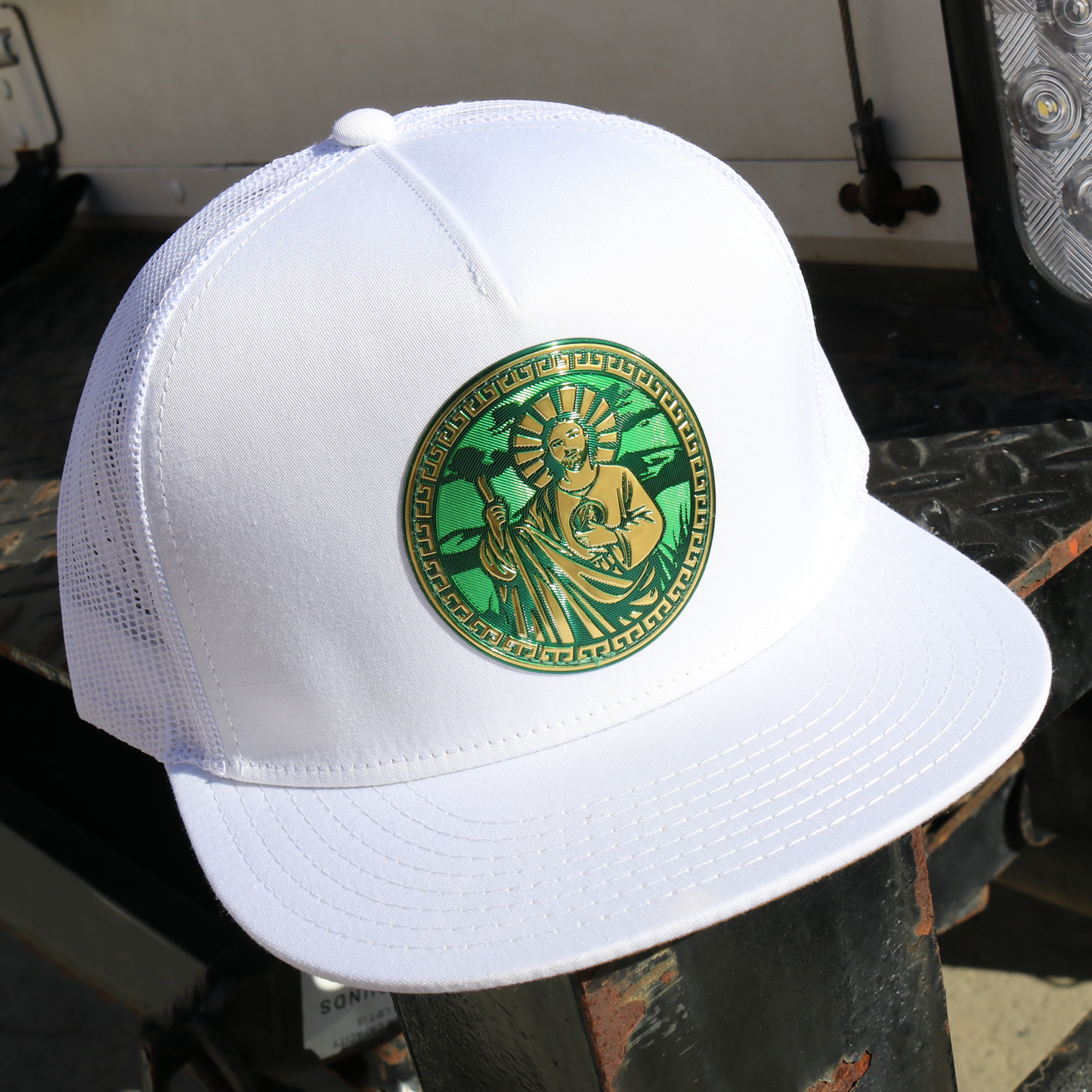 San Judas white flat visor trucker hat with green and gold circular emblem.