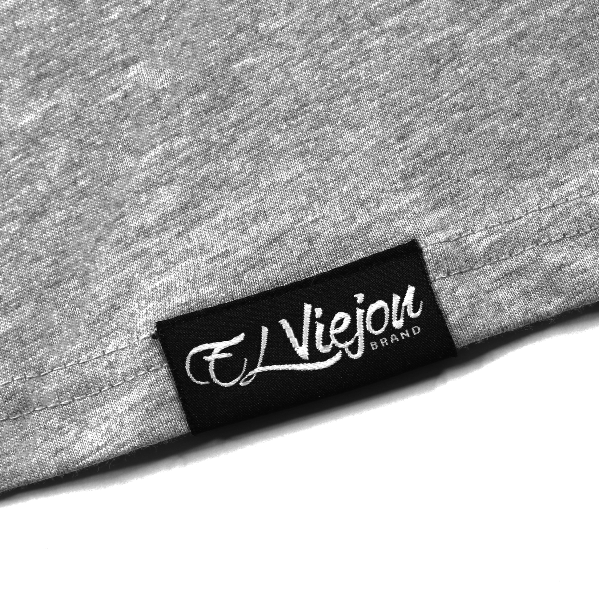 Close-up of a heather gray Alpha Shirt's hem with a black "El Viejon Brand" woven label.