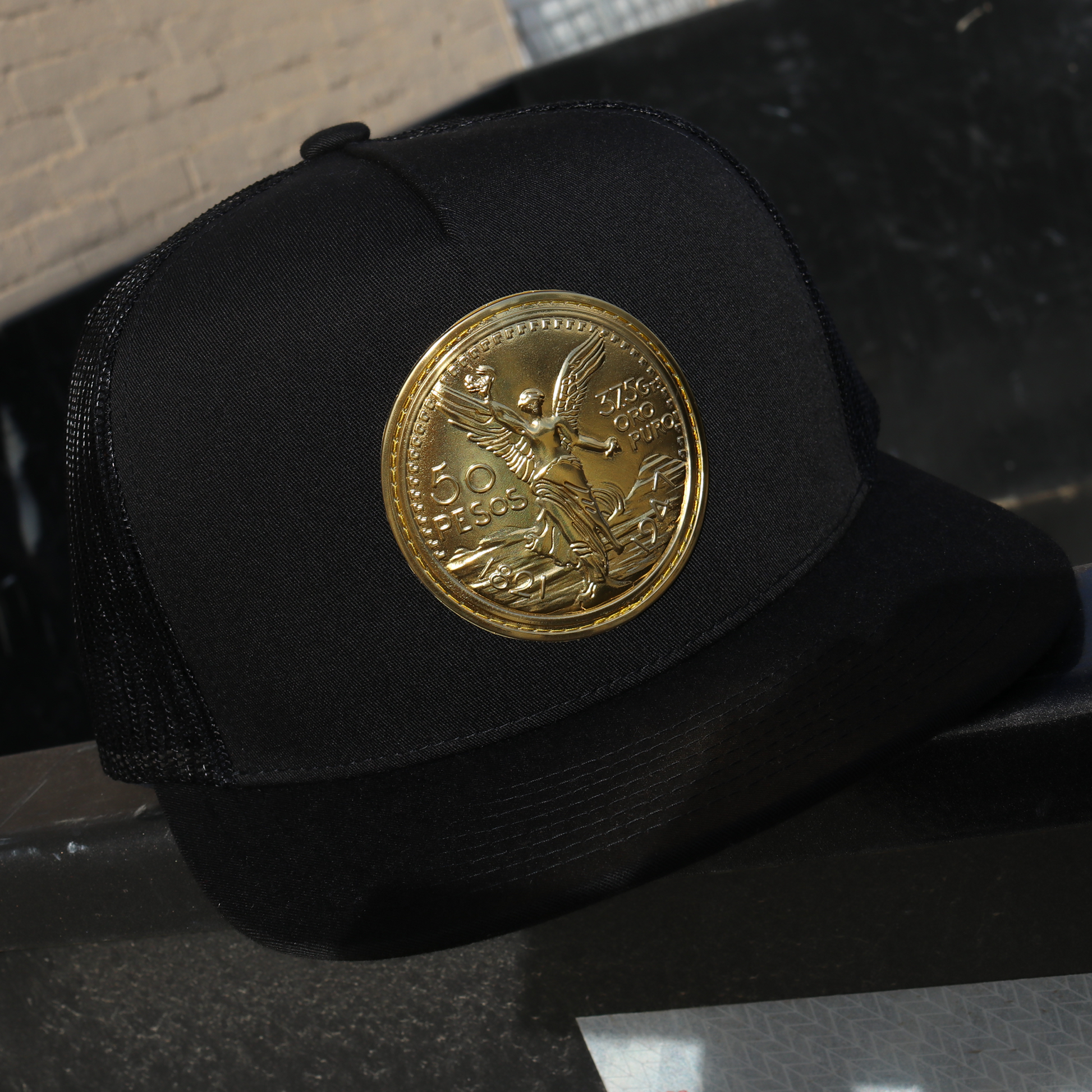 Black trucker hat with flat visor, black mesh, and a gold El Cente coin emblem.