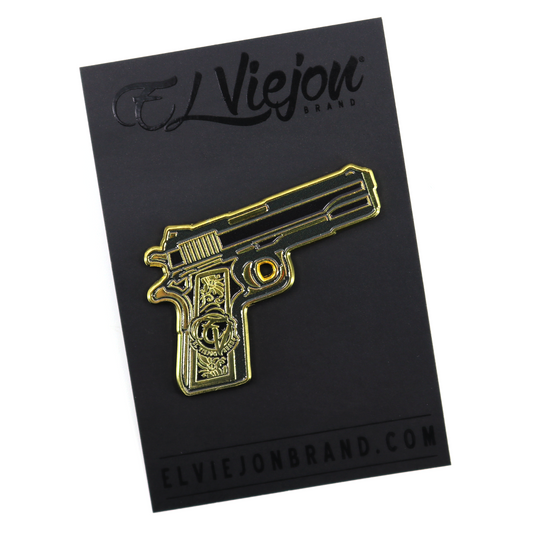 Pin dorado de pistola con cachas de oro y detalles grabados, sobre tarjeta negra El Viejon Brand.