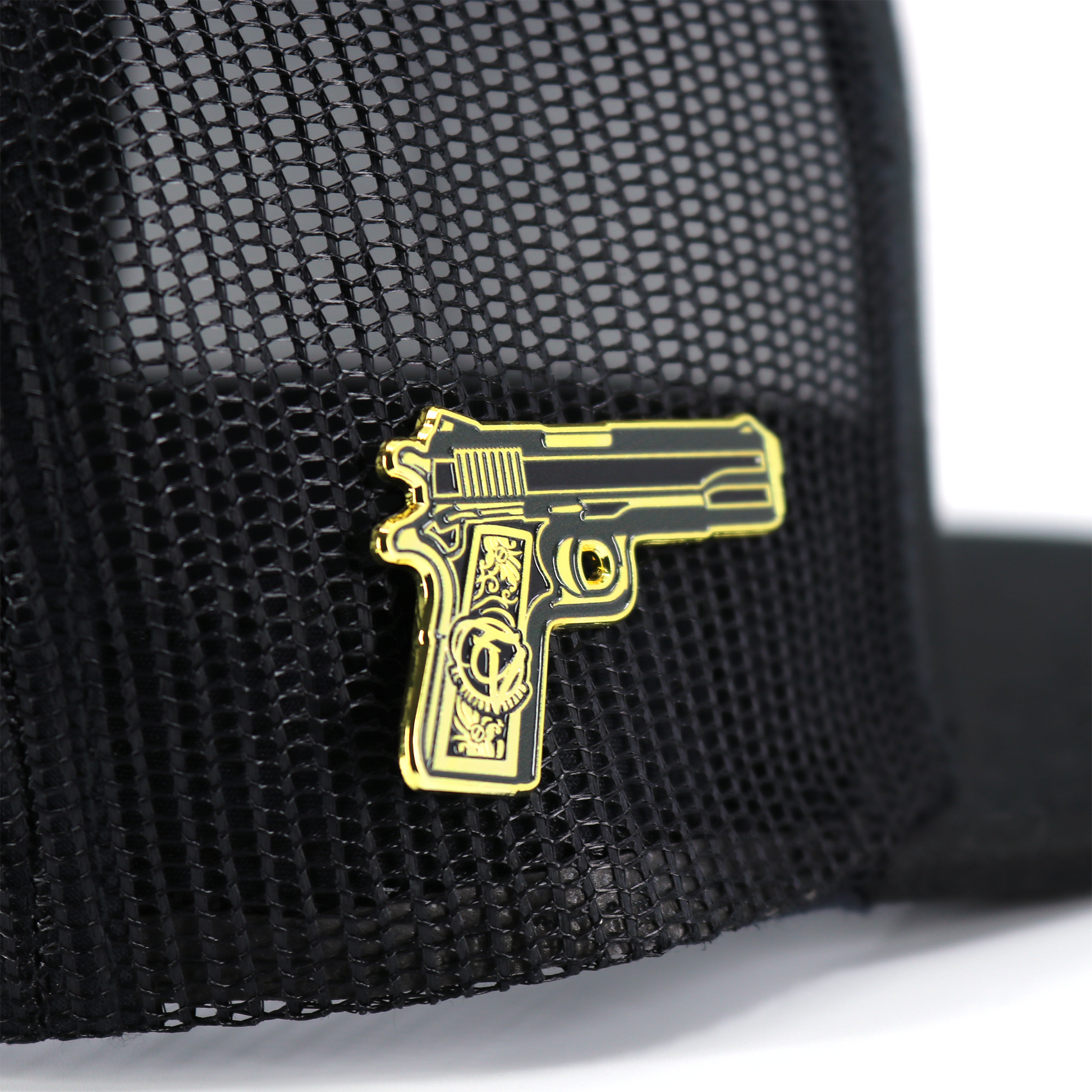 Pin de pistola dorada y negra con diseño detallado, sujetado a una gorra de malla negra.