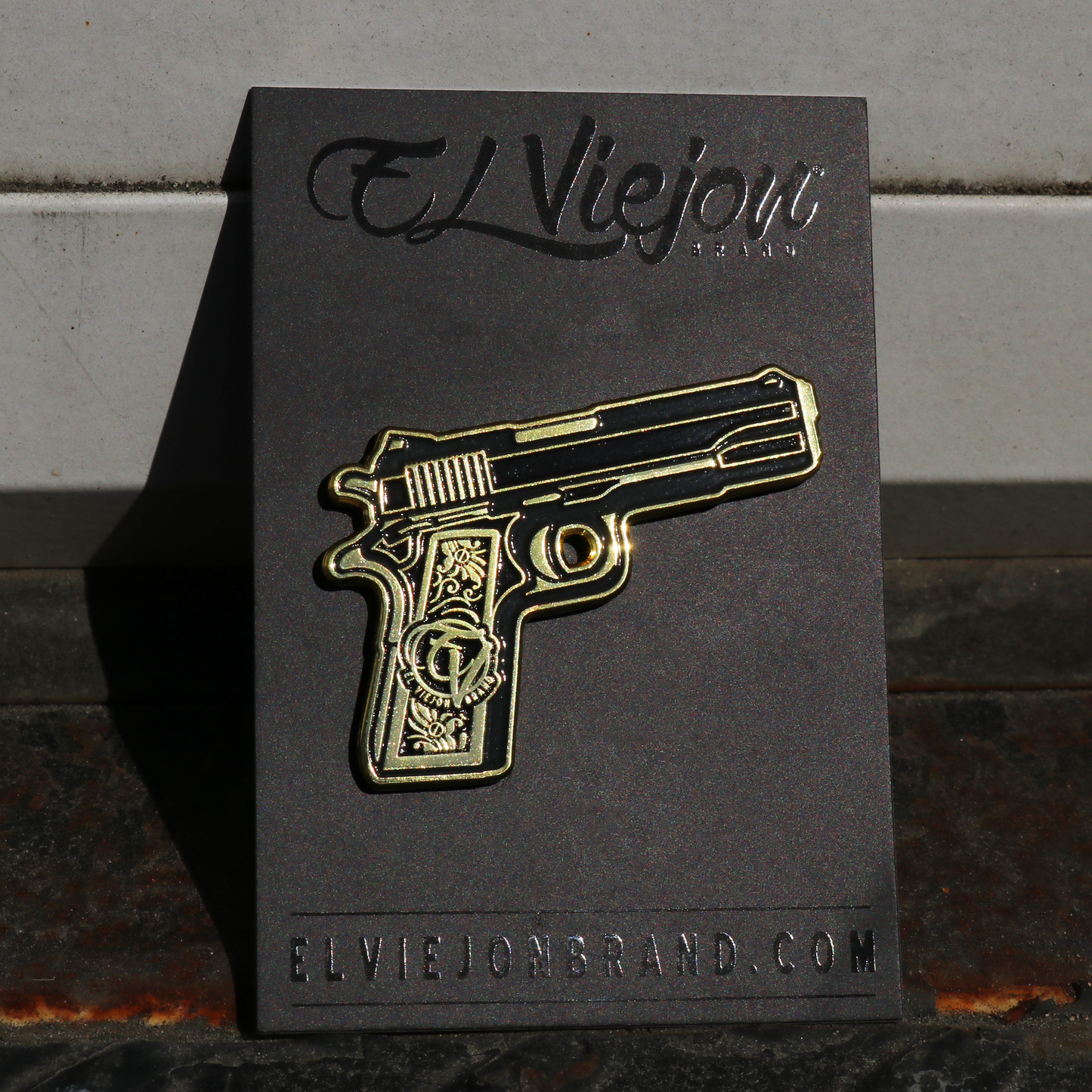 Pin de pistola dorada y negra sobre tarjeta negra El Viejon Brand.