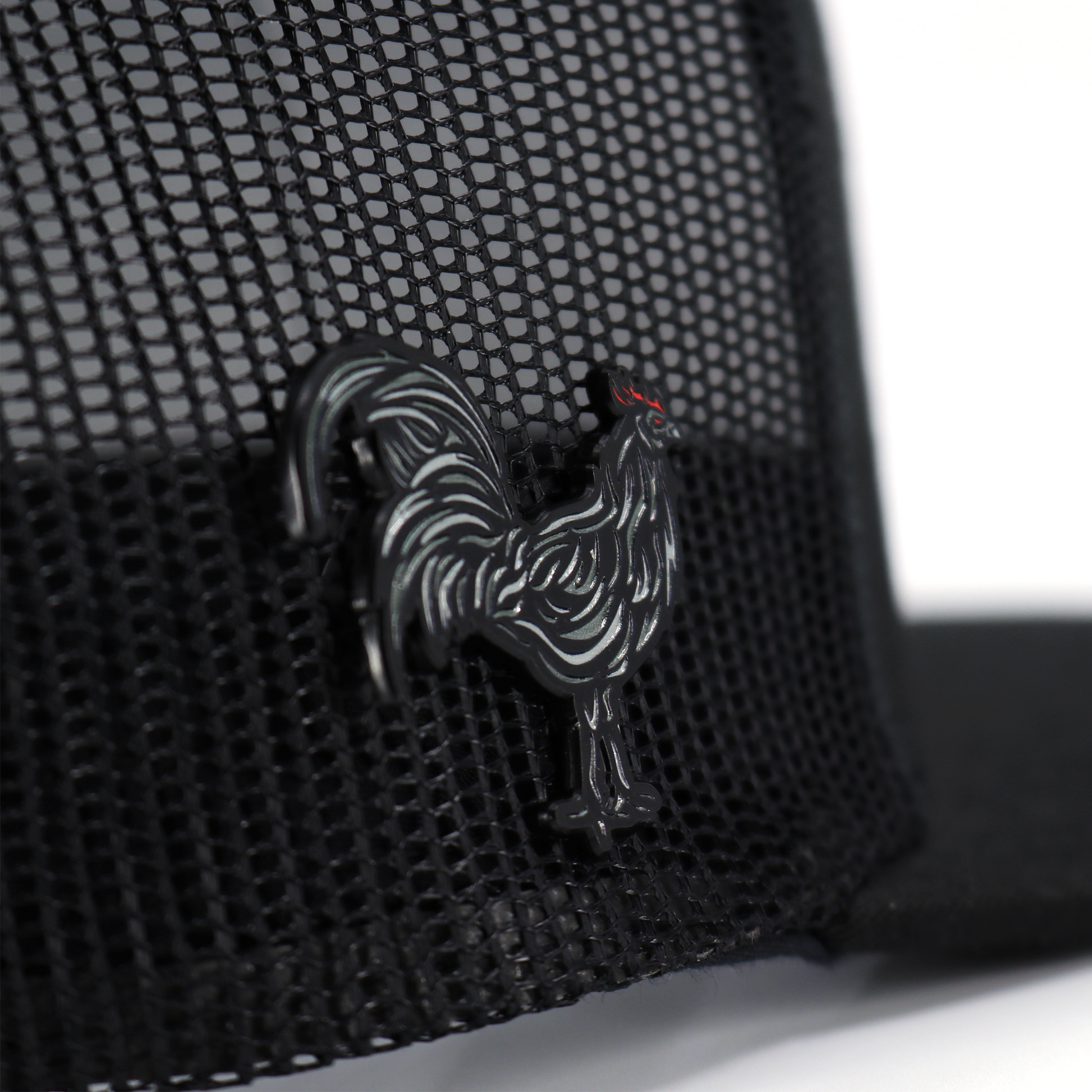 Black Gallo Negro enamel pin with red comb, attached to a black mesh hat.