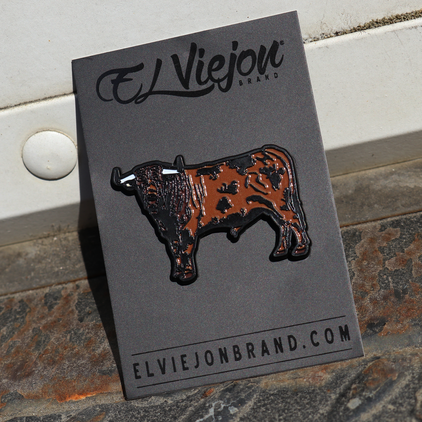Pin esmaltado de toro semental marrón y negro con cuernos blancos, en tarjeta negra "El Viejon Brand".