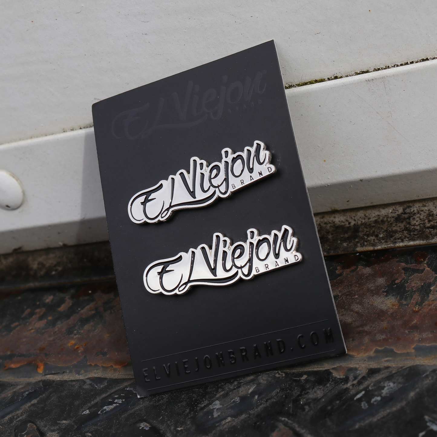 Two silver El Viejon Brand pins with black outlines on a black card.