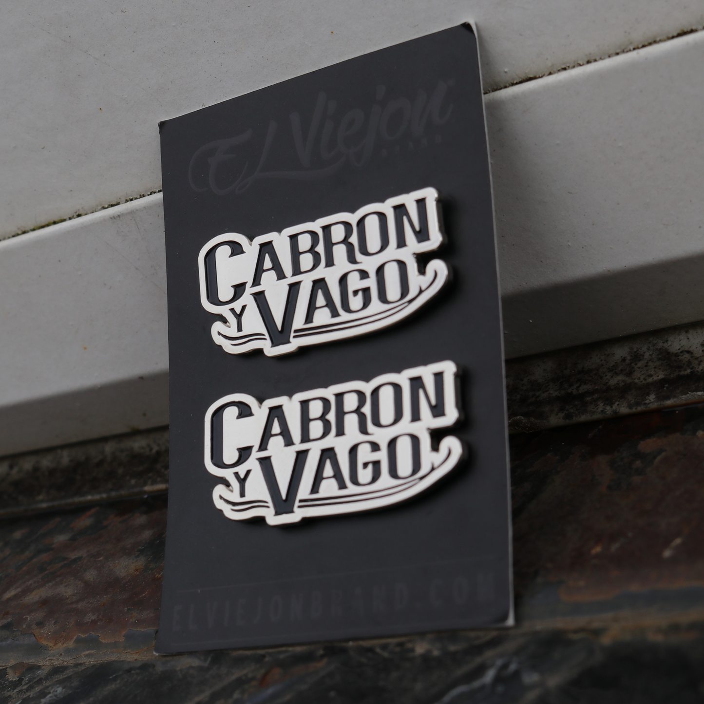 Two black and white "Cabron y Vago" enamel pins on a dark gray backing card.