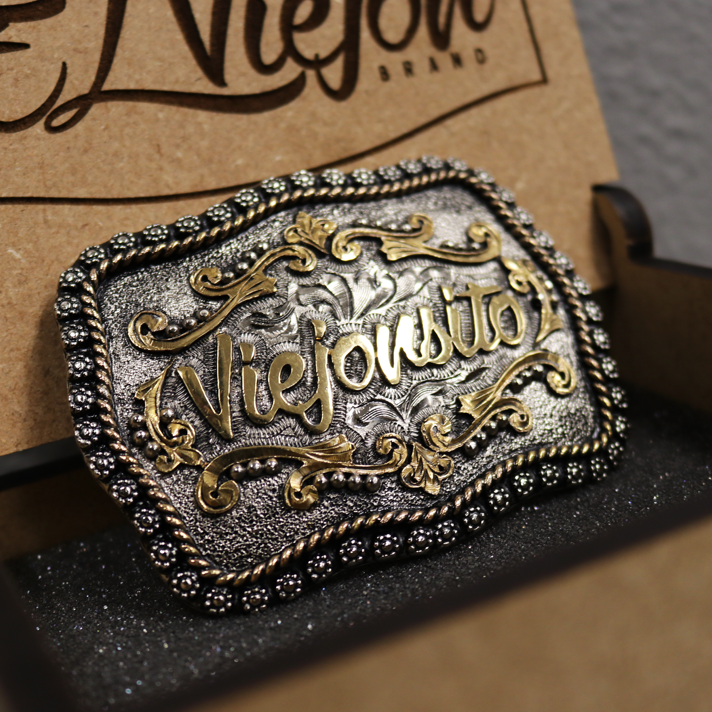 Hebilla artesanal "Viejonsito" para niño, de metal plateado con letras doradas y detalles grabados, en caja de madera.
