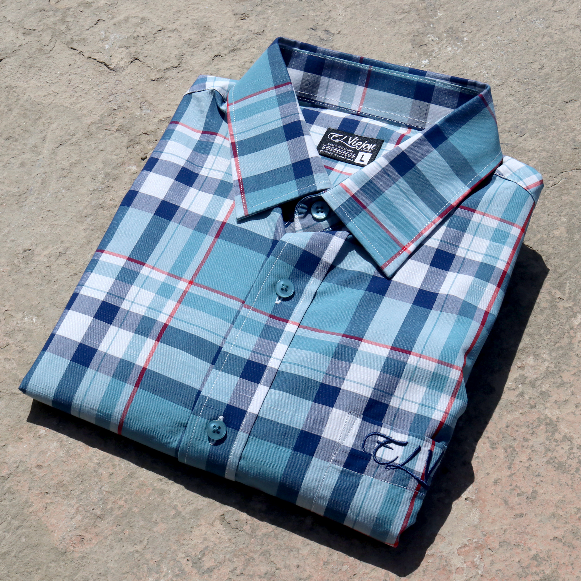 Men's plaid dress shirt in teal, navy, and red, folded on a concrete surface. Features a straight collar and front pocket with embroidered logo.