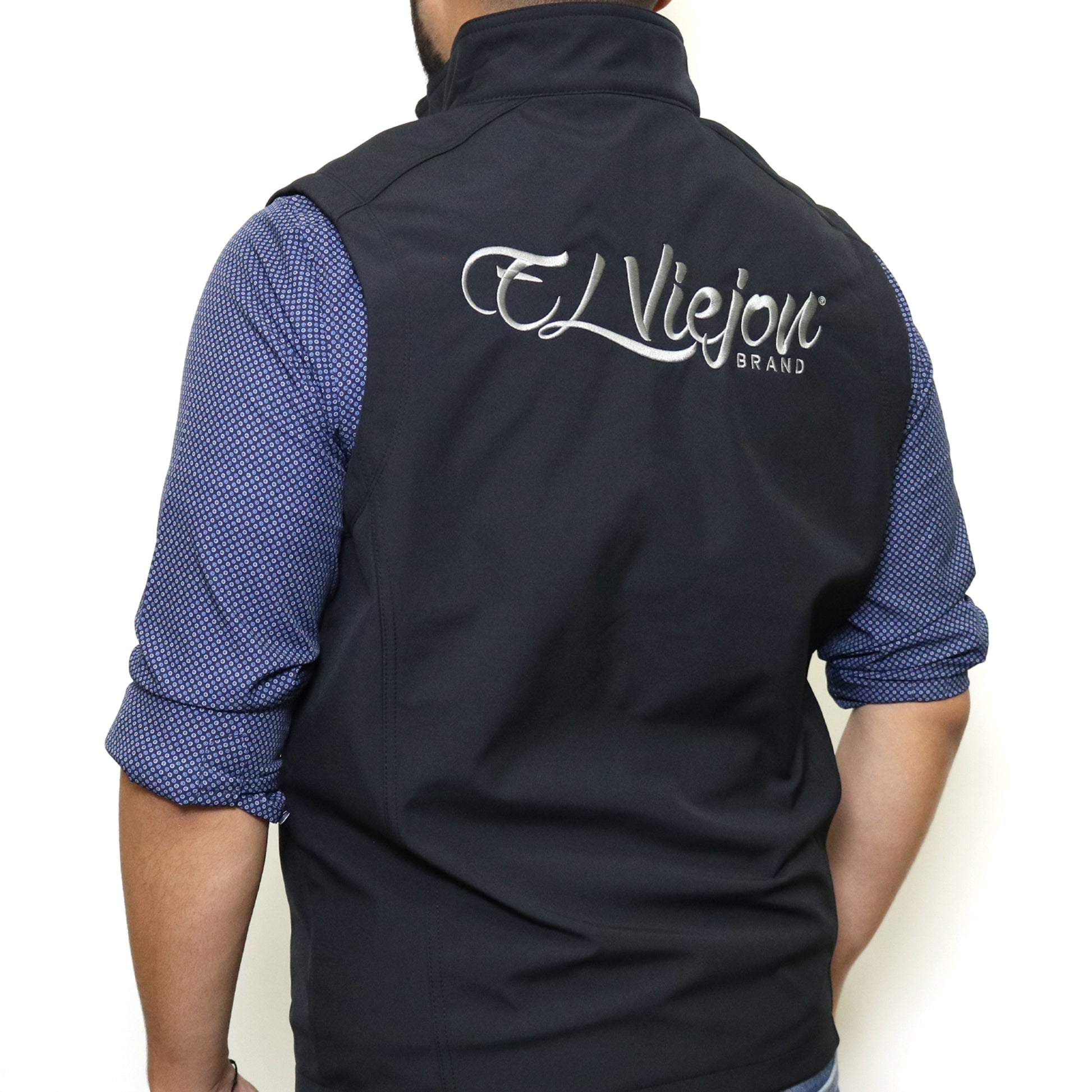 Black El Viejon Brand Vest, back view, with charcoal embroidered logo, worn over a blue patterned shirt.