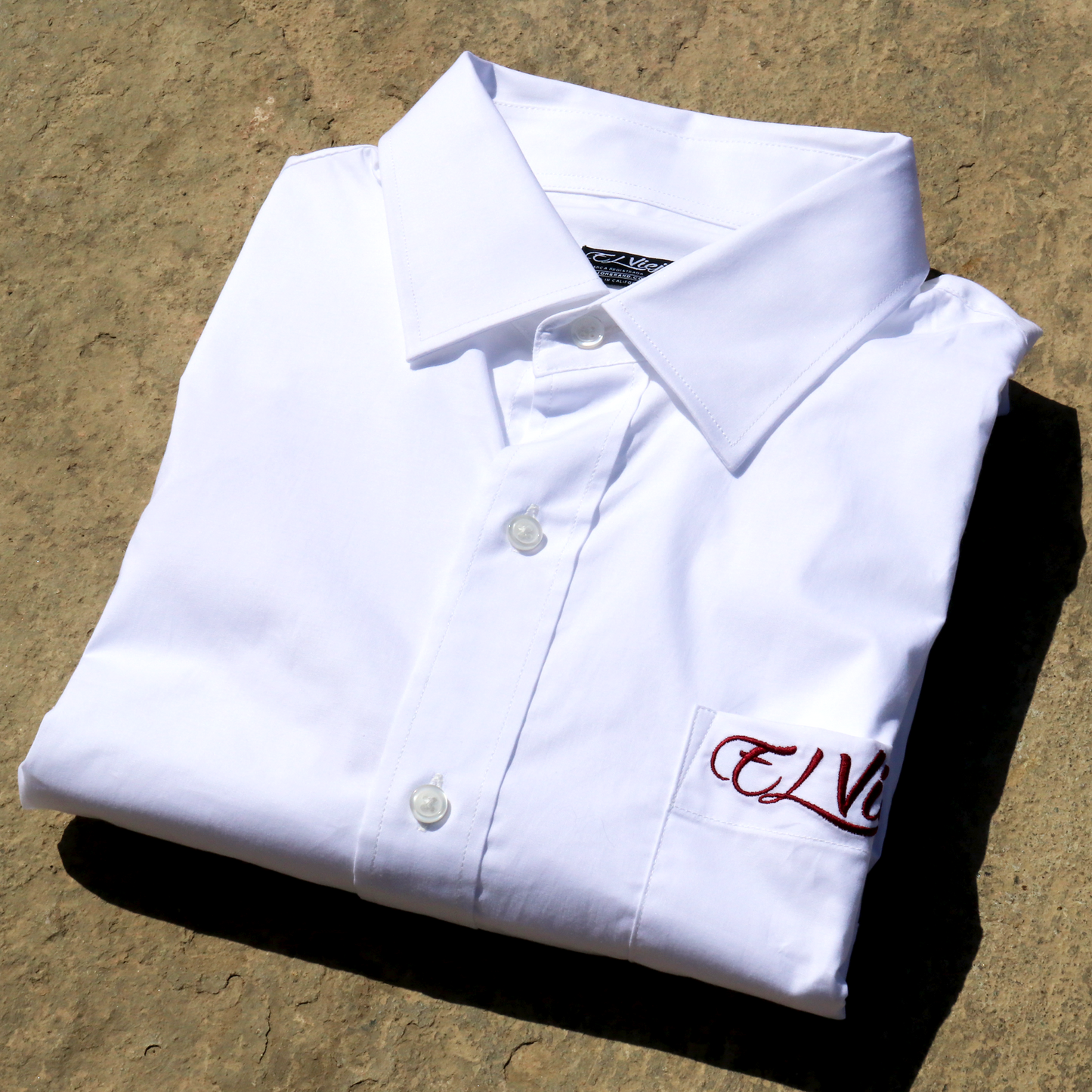 Folded white cotton-span dress shirt with straight collar, front pocket, and red "El Viejon Brand" embroidered logo.
