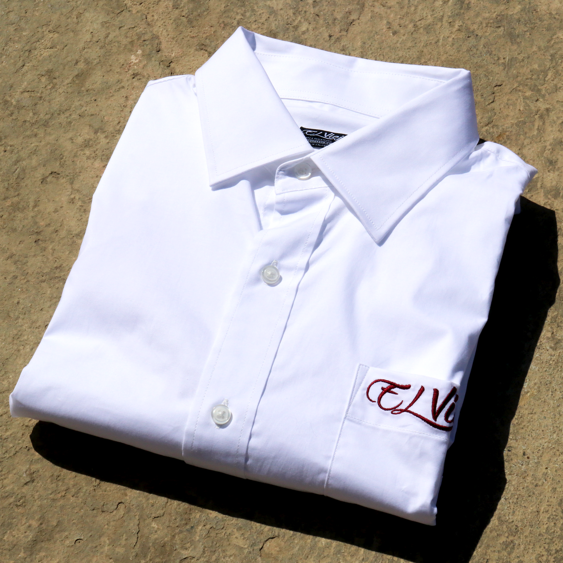 Folded white cotton-span dress shirt with straight collar, front pocket, and red "El Viejon Brand" embroidered logo.